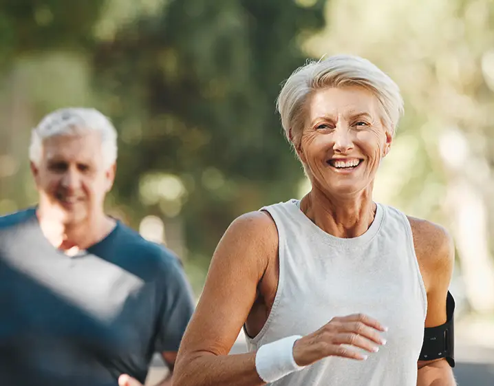 An older couple smiling as they run through a forest.